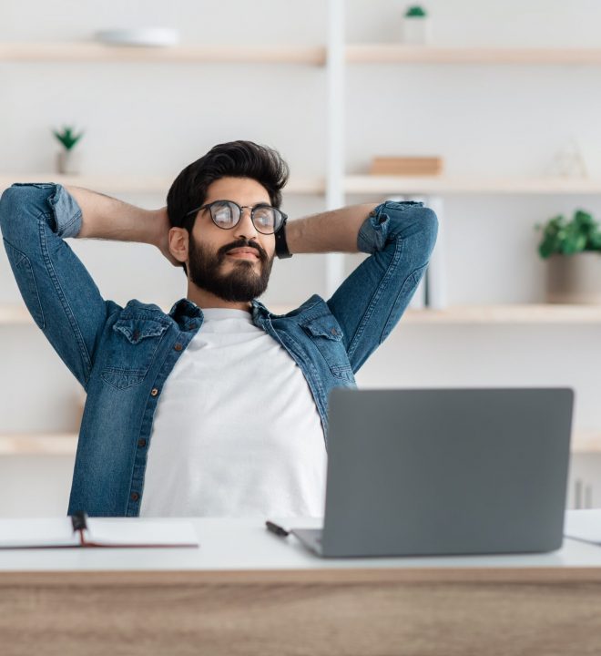 Successful arab freelancer guy resting holding hands behind head, relaxing at workplace after online work, sitting at desk at home office, empty space